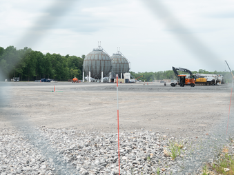 Two giant gray hydrogen plant storage tanks are framed by the diamond of a chain link fence. Forest is to the left of the globes, large machinery to the right, and barren gravel between the globes and the fence.