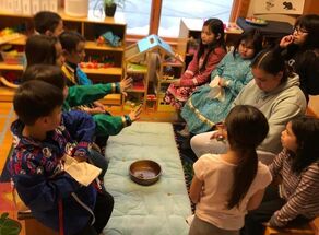 A group of children sits on a rug with animals on it around a long floor pillow with a bowl on it. They are in a classroom, and behind them is a window, a dollhouse, and shelves with other classroom items.