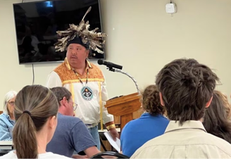 Tonawanda Chief Roger Hill dressed in traditional dress speaks into a microphone at a podium. Behind him is a large black flatscreen TV and in the foreground are the backs of the heads of people in the audience who are facing him.